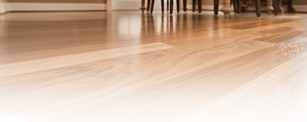 Close-up of a polished wooden floor with varying shades of brown, reflecting light. The background shows a dining table and chairs, partially visible at the edge of the room, with soft natural light coming through a window.