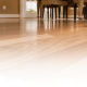 Close-up of a polished wooden floor with varying shades of brown, reflecting light. The background shows a dining table and chairs, partially visible at the edge of the room, with soft natural light coming through a window.